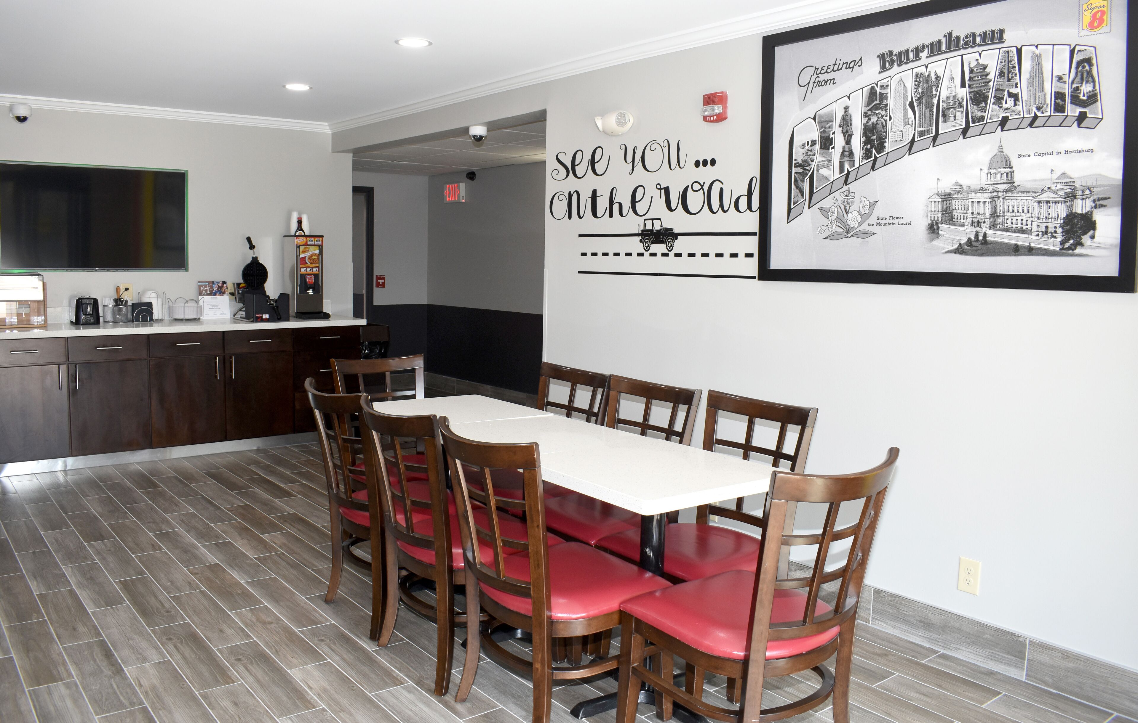 Un comedor con una mesa larga y sillas, una cocina al fondo y una pared con un cartel de bienvenida.