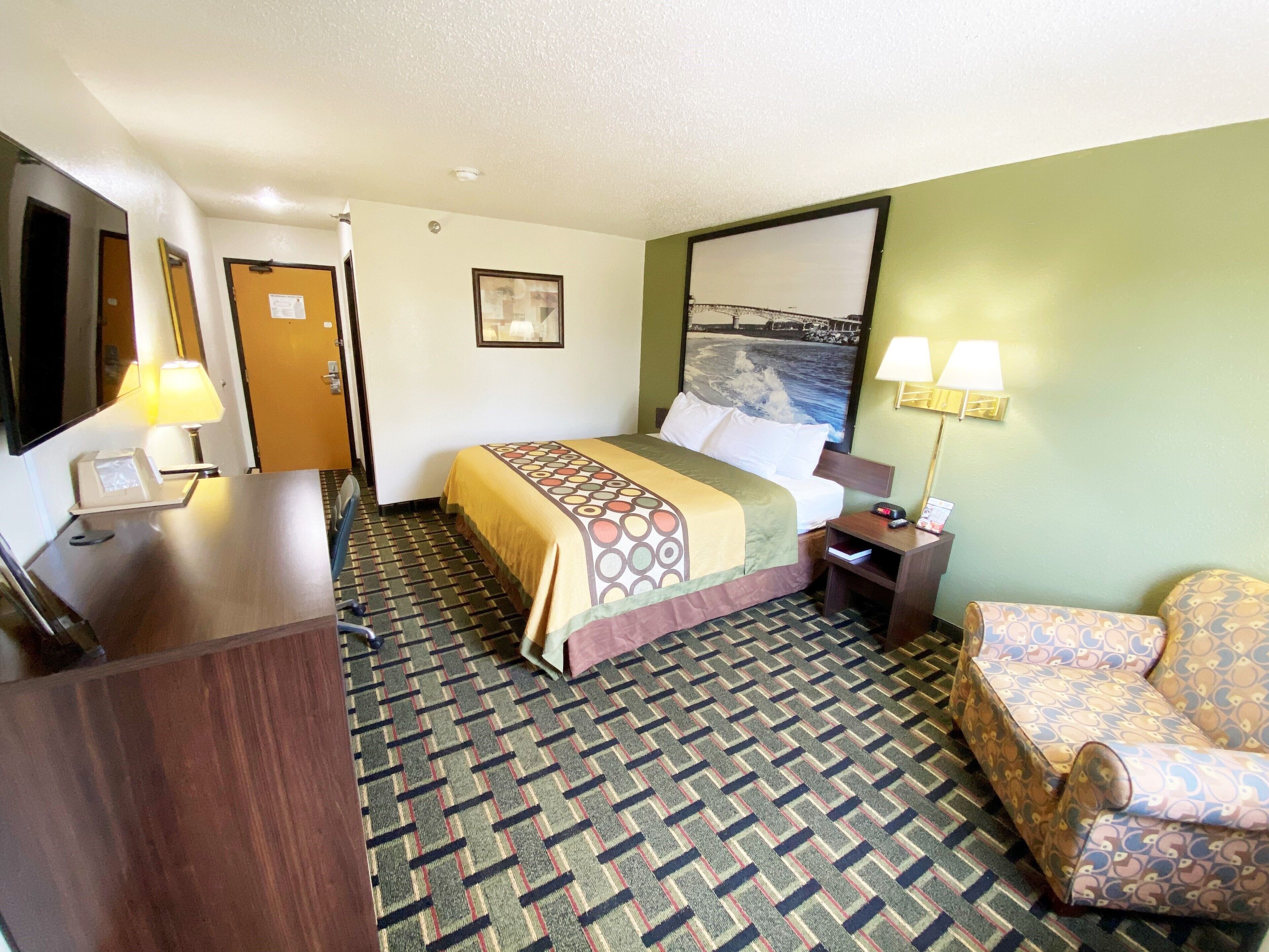 Quarto de hotel com cama, escrivaninha, cadeira, luminária, quadro e tapete estampado.
