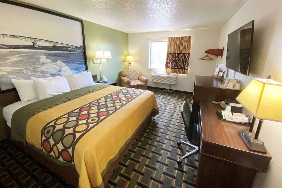 A hotel room with a bed, a desk, a chair, a window with curtains, a painting of a bridge, and a wall-mounted TV.