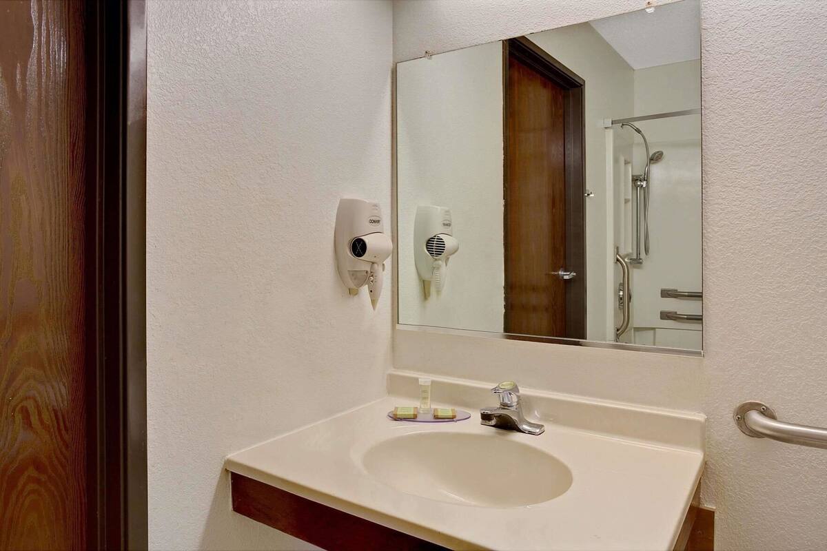 A bathroom with a white sink, a mirror, a hairdryer, and a shower visible in the reflection.