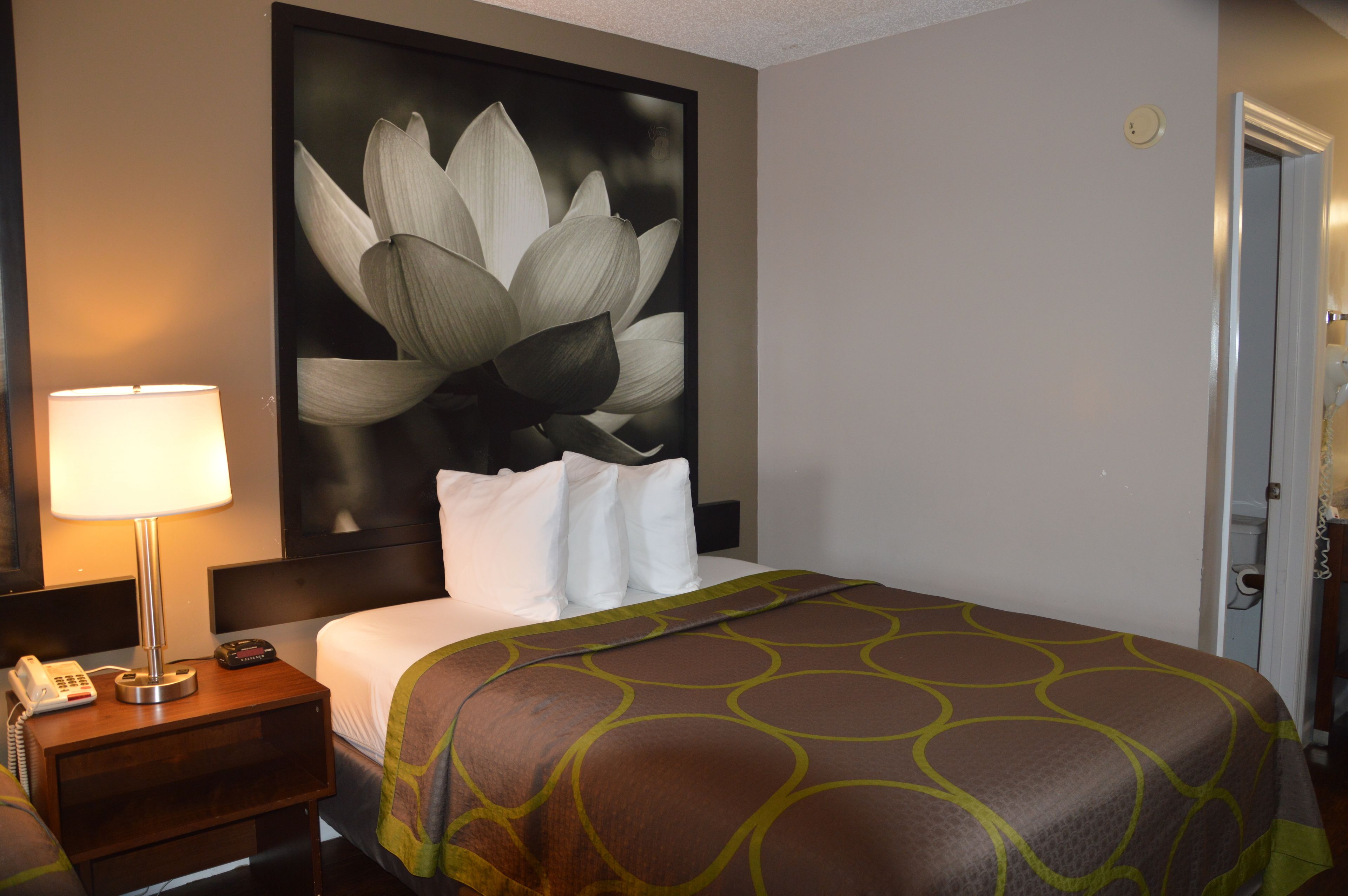 A hotel room with a large bed, a nightstand with a lamp, a telephone, and a wall-mounted artwork of a flower.