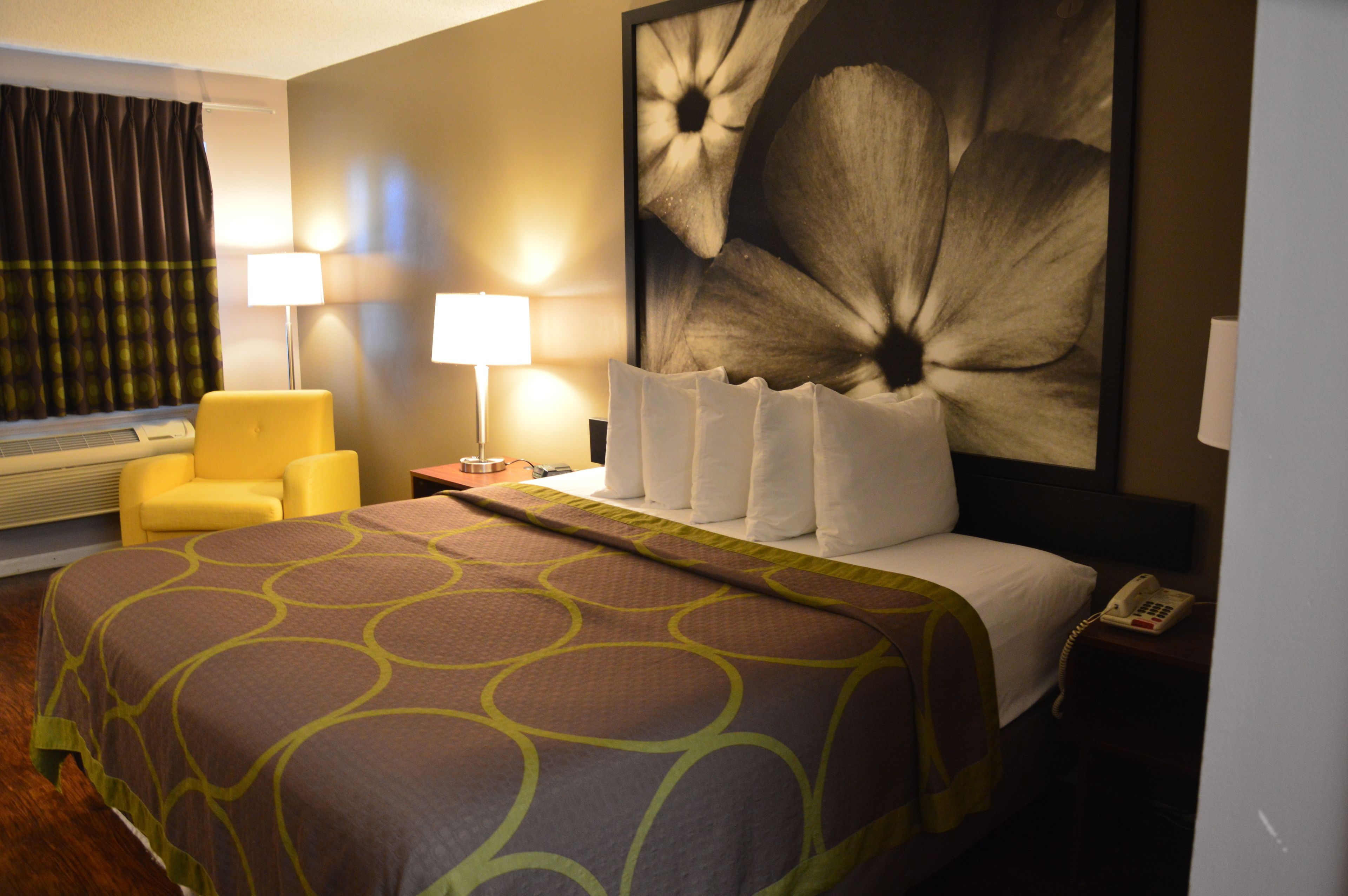 Una habitación de hotel con una cama grande, un sillón amarillo, dos lámparas de noche y un cuadro floral grande en la pared.