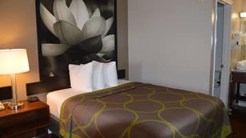 A hotel room with a large bed, a nightstand with a lamp, a wall-mounted artwork of flowers, and a door with a mirror and towel rack.