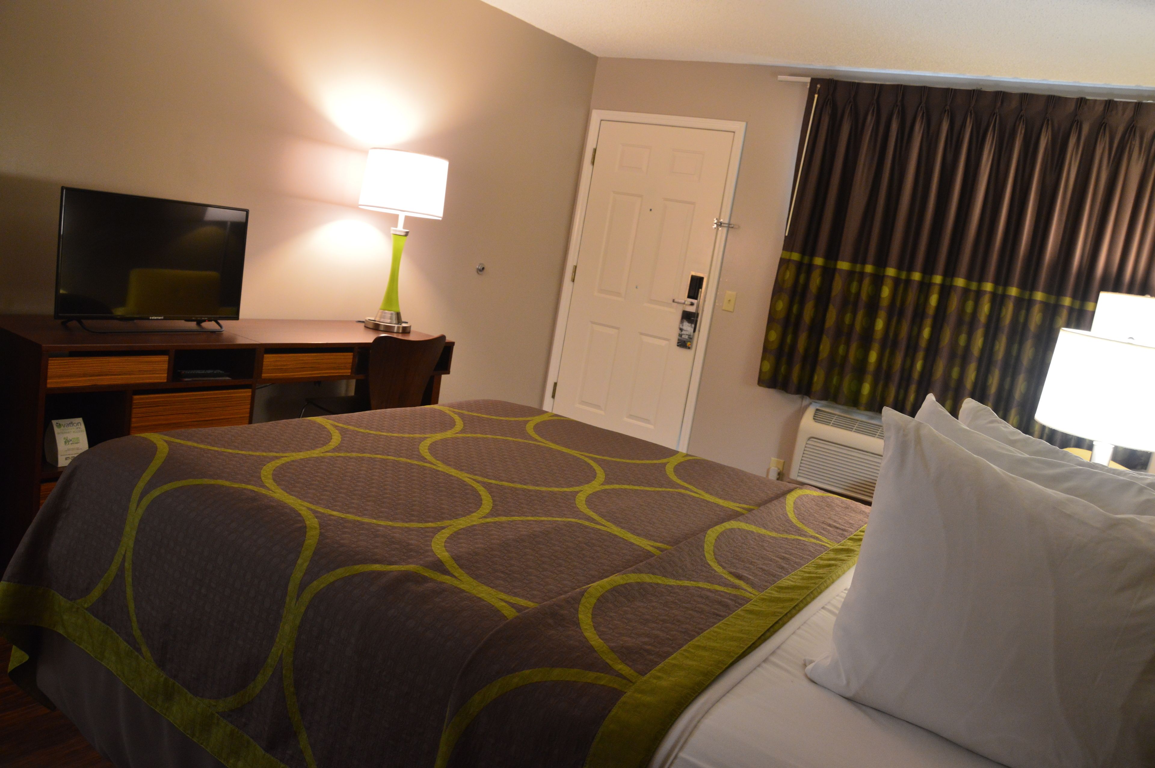 A hotel room with a bed, a desk with a television, a lamp, a door, and an air conditioning unit.