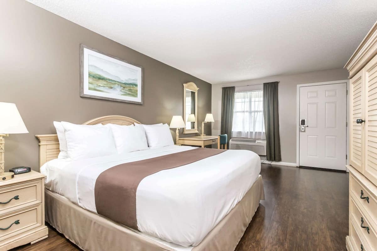 A hotel room with a large bed, bedside tables, a desk, a chair, a mirror, a wardrobe, and a painting on the wall.