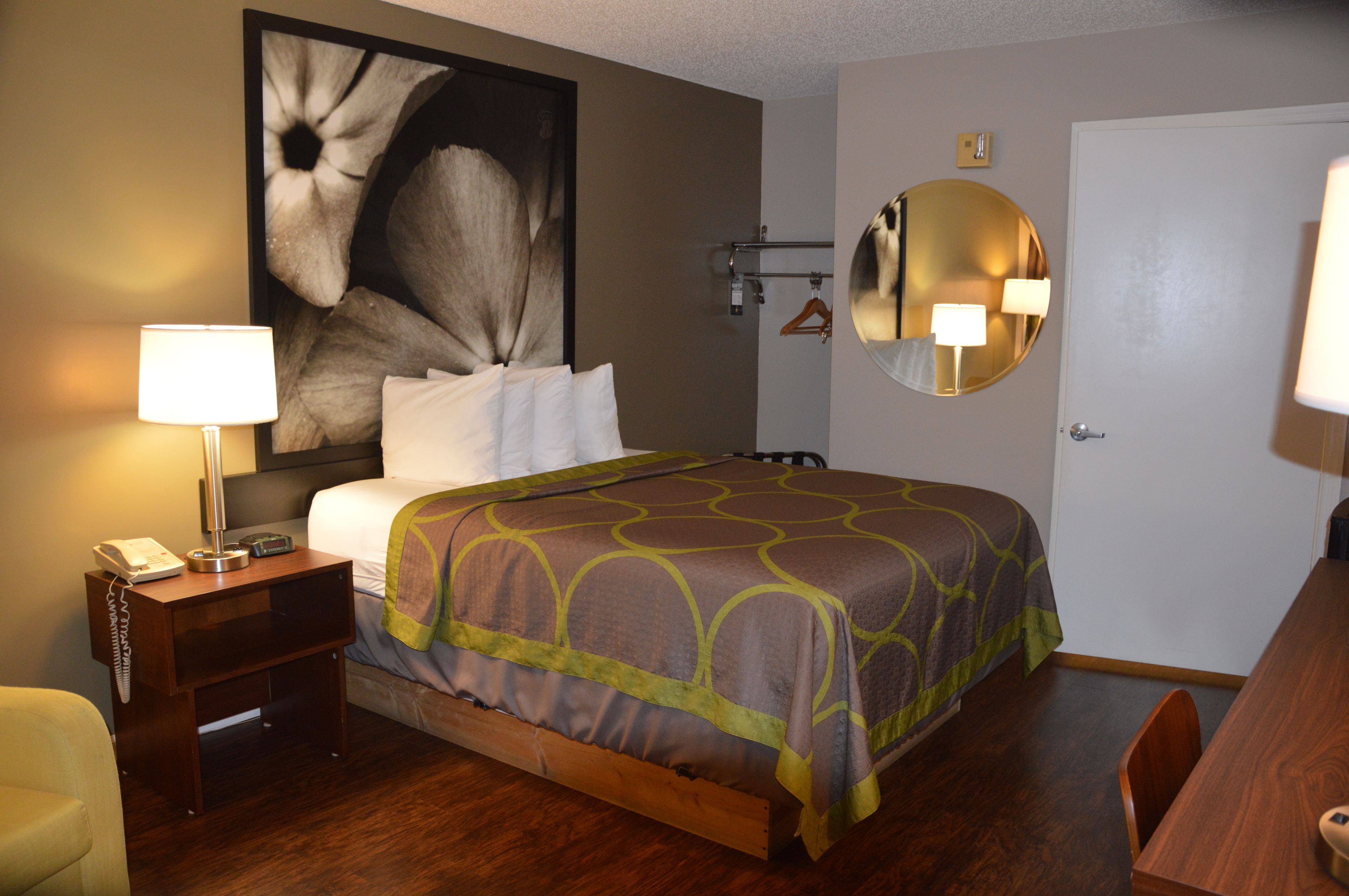 A hotel room with a bed, bedside table with a lamp, a chair, a wooden table, a round mirror, and a large floral artwork on the wall.
