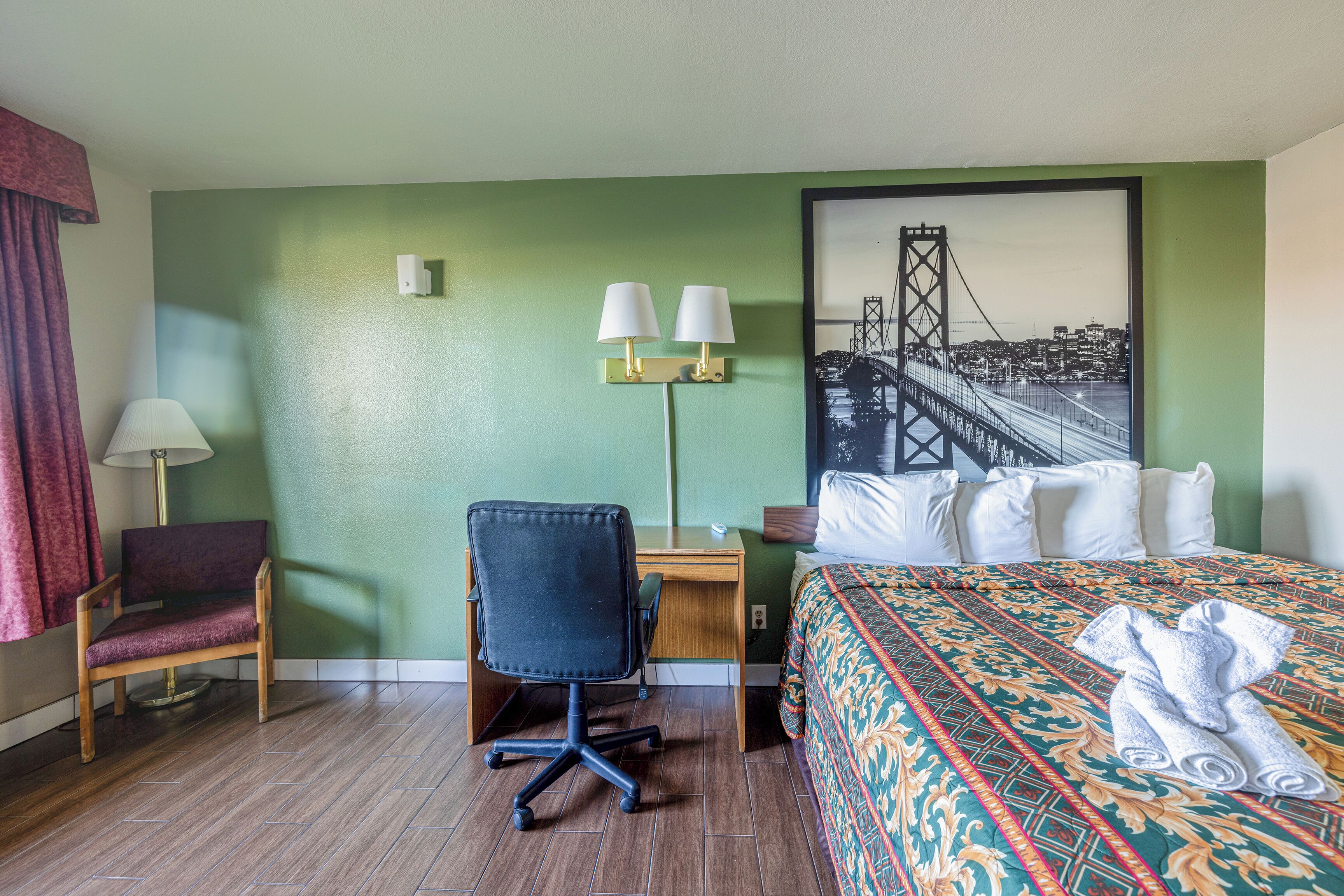 A hotel room with a bed, a desk, a chair, a lamp, and a cityscape painting on the wall.