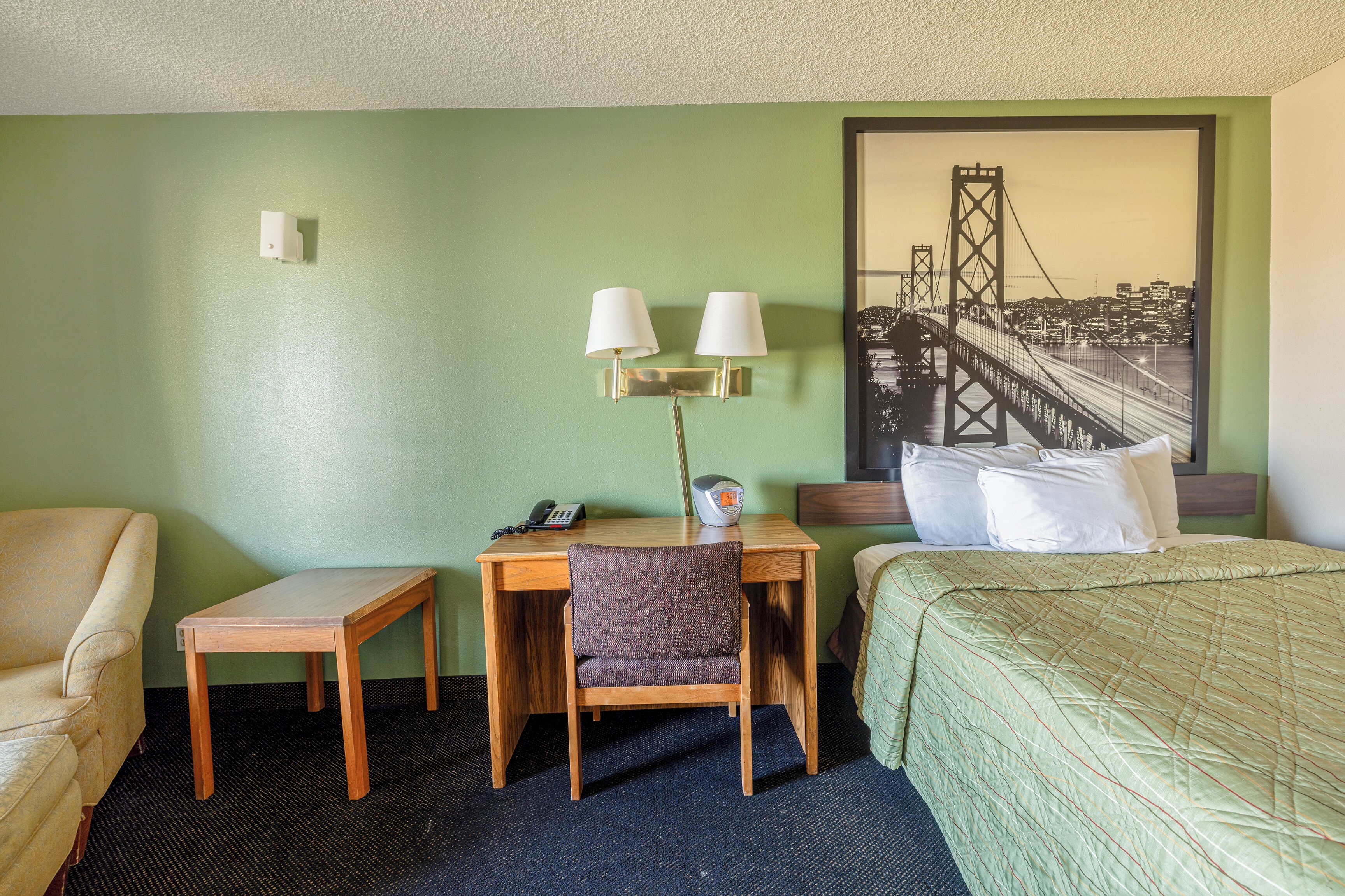 A hotel room with a desk, chair, bed, and a painting of a bridge.