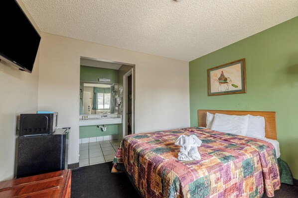 Habitación de hotel con cama, televisor, refrigerador pequeño, baño con lavamanos y espejo, y pared verde con un cuadro enmarcado.