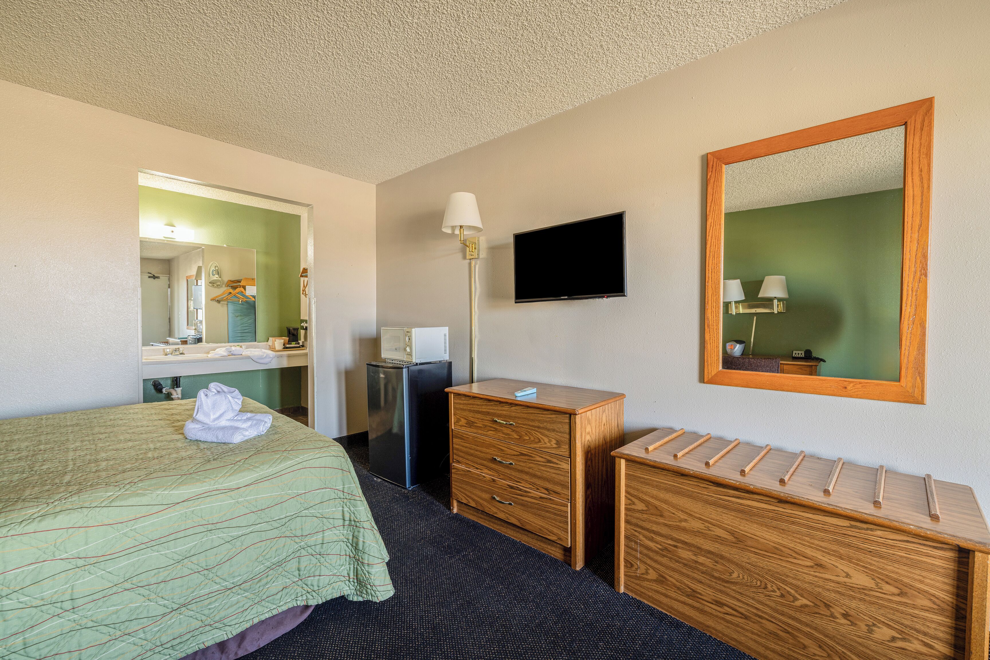 A hotel room with a bed, a wooden dresser, a desk, a mirror, a television, and a lamp.