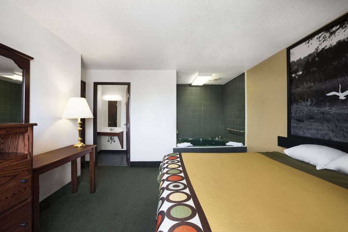 Ein Hotelzimmer mit einem Bett, einem Schreibtisch mit Lampe, einem Badezimmer mit Badewanne und einem großen gerahmten Bild an der Wand.