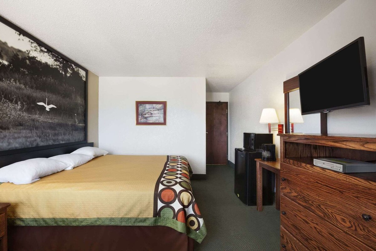 Ein Hotelzimmer mit einem Bett, einem Fernseher, einem Schreibtisch mit Lampe und einem Gemälde an der Wand.