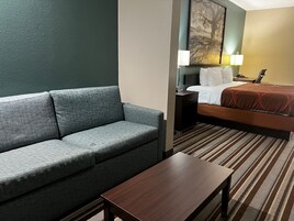 Habitación de hotel con un sofá gris, una mesa de madera, una cama con colcha marrón, una mesita de noche con lámpara y un cuadro en la pared.