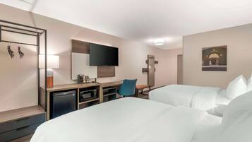 A hotel room with two beds, a desk, a chair, a television, and a small kitchenette.
