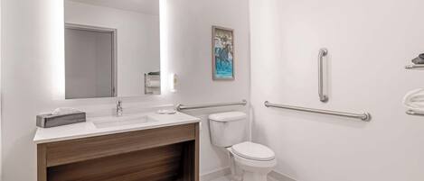 Un baño moderno con lavamanos, espejo y retrete blancos. Hay barras de apoyo en las paredes.