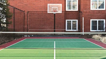 A tennis court with a net, a basketball hoop, and a brick building in the background.