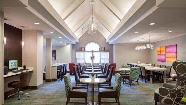 A hotel dining area with multiple tables and chairs, a central chandelier, and a view of the lobby through a large arched window. - Cambridge Suites Mississauga (Mississauga)