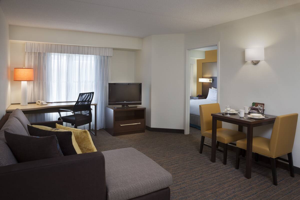 A hotel room with a sofa, a TV, a desk with a chair, and a dining table with chairs.
