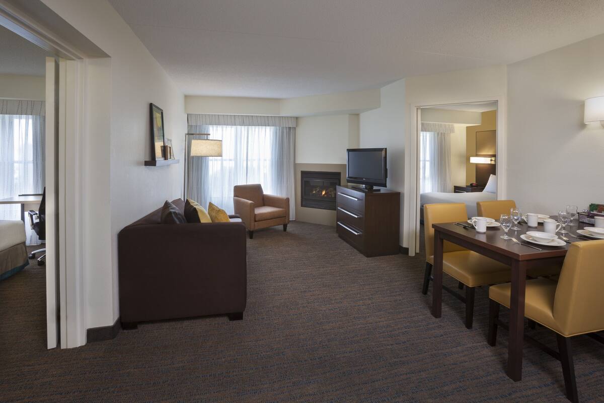 A hotel room with a living area, a dining table set for two, a sofa, a television, and a fireplace.