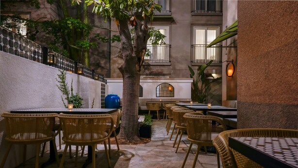 An outdoor patio with wicker tables and chairs, stone flooring, and a view of a building with balconies. - Hotel Amarano Burbank - Hollywood (Burbank)