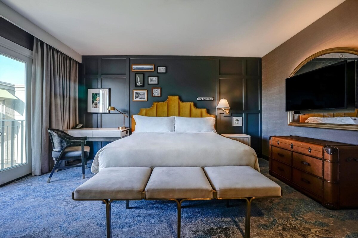 Une chambre d’hôtel avec un grand lit, un bureau, une chaise, un banc, une commode et un téléviseur.