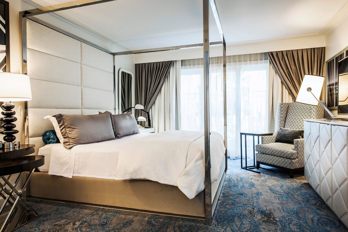 Une chambre d’hôtel avec un lit, une chaise, un bureau, une lampe et une grande fenêtre avec des rideaux.
