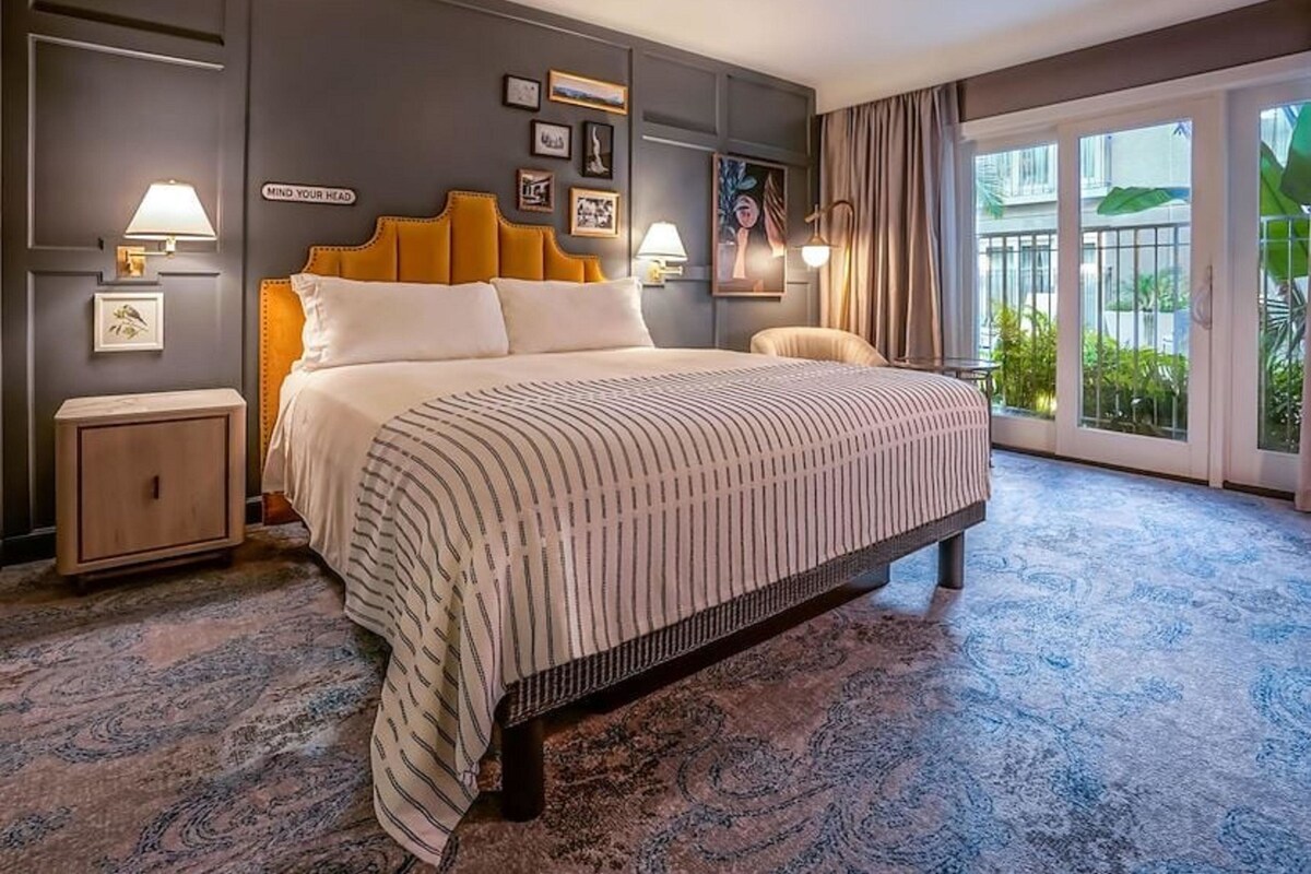 Une chambre d’hôtel avec un grand lit, une table de chevet, une chaise, une vue sur l’extérieur et un mur orné de tableaux et d’une affiche.