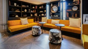 A modern lounge area with a yellow sofa set, round coffee tables, and a dark bookshelf filled with books and decorative items. - Hotel Amarano Burbank - Hollywood (Burbank)