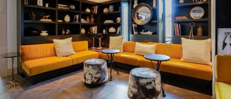 Un área de descanso moderna con un conjunto de sofá amarillo, mesas de centro redondas y una estantería oscura llena de libros y objetos decorativos.