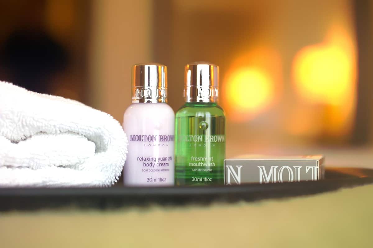 Twee flessen Molton Brown body cream en mondwater, een opgevouwen witte handdoek, en een bord met het Molton Brown logo.