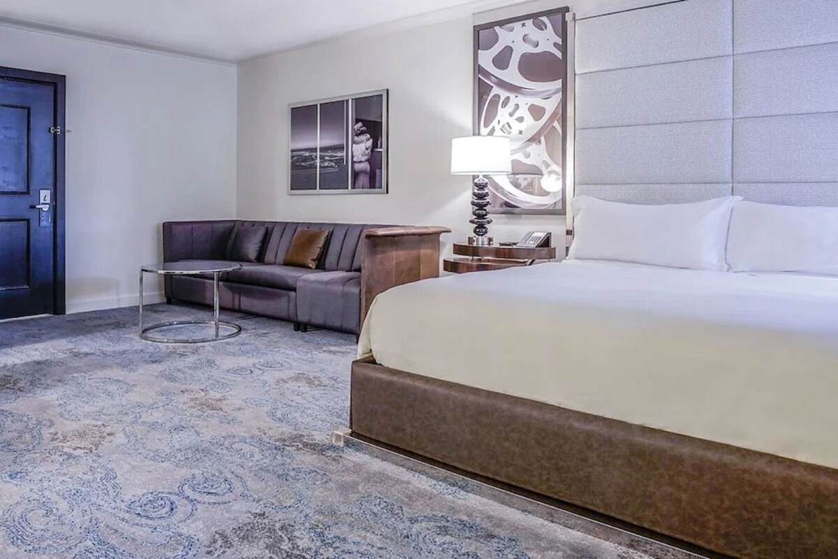 Une chambre d’hôtel avec un grand lit, un canapé, une table de chevet avec une lampe, une œuvre d’art accrochée au mur et une porte.