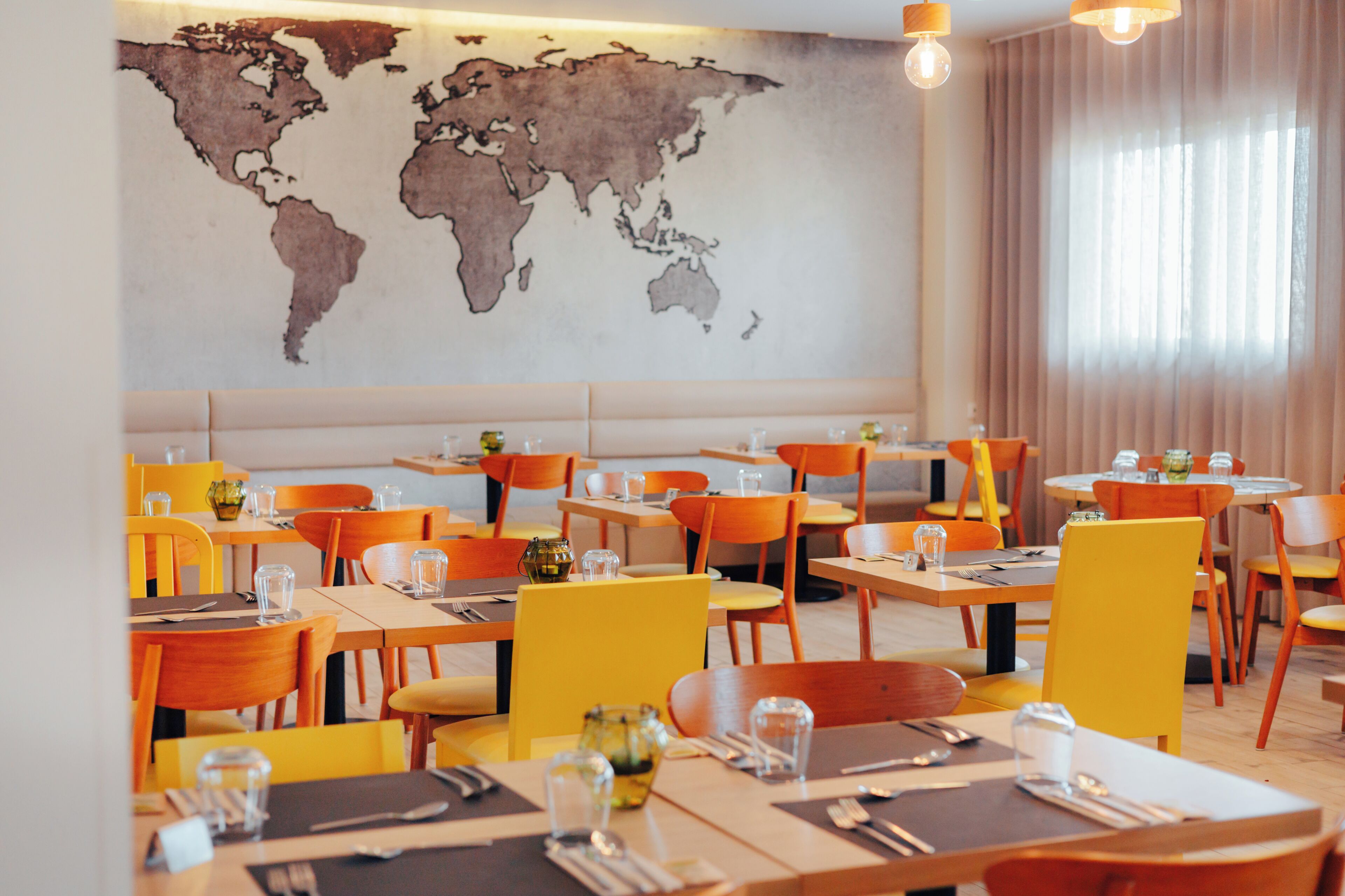 A restaurant with multiple tables and chairs, a world map on the wall, and a large window with curtains.