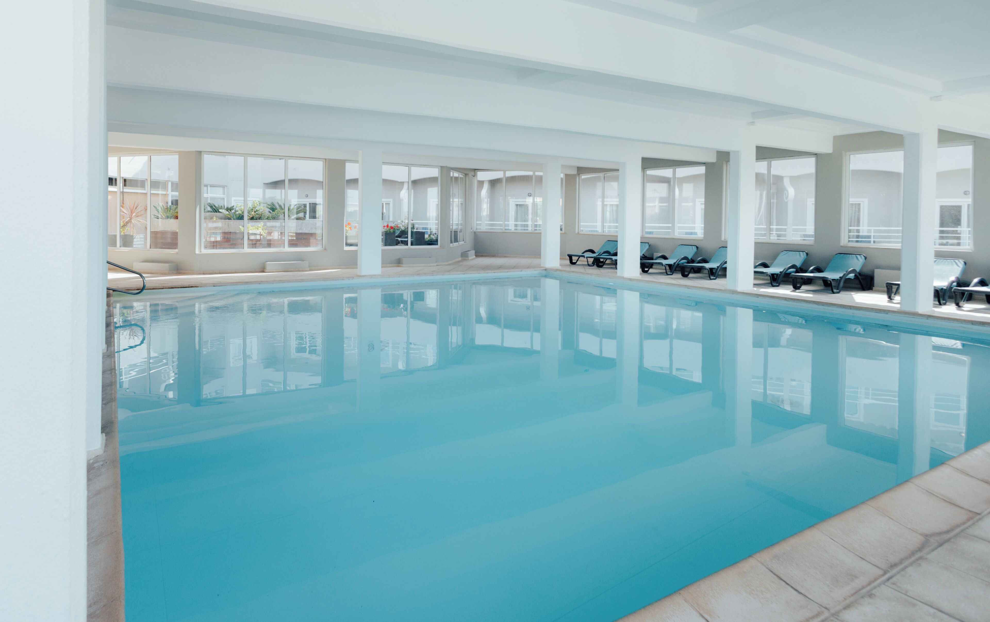 An indoor swimming pool with clear blue water, surrounded by lounge chairs and large windows.
