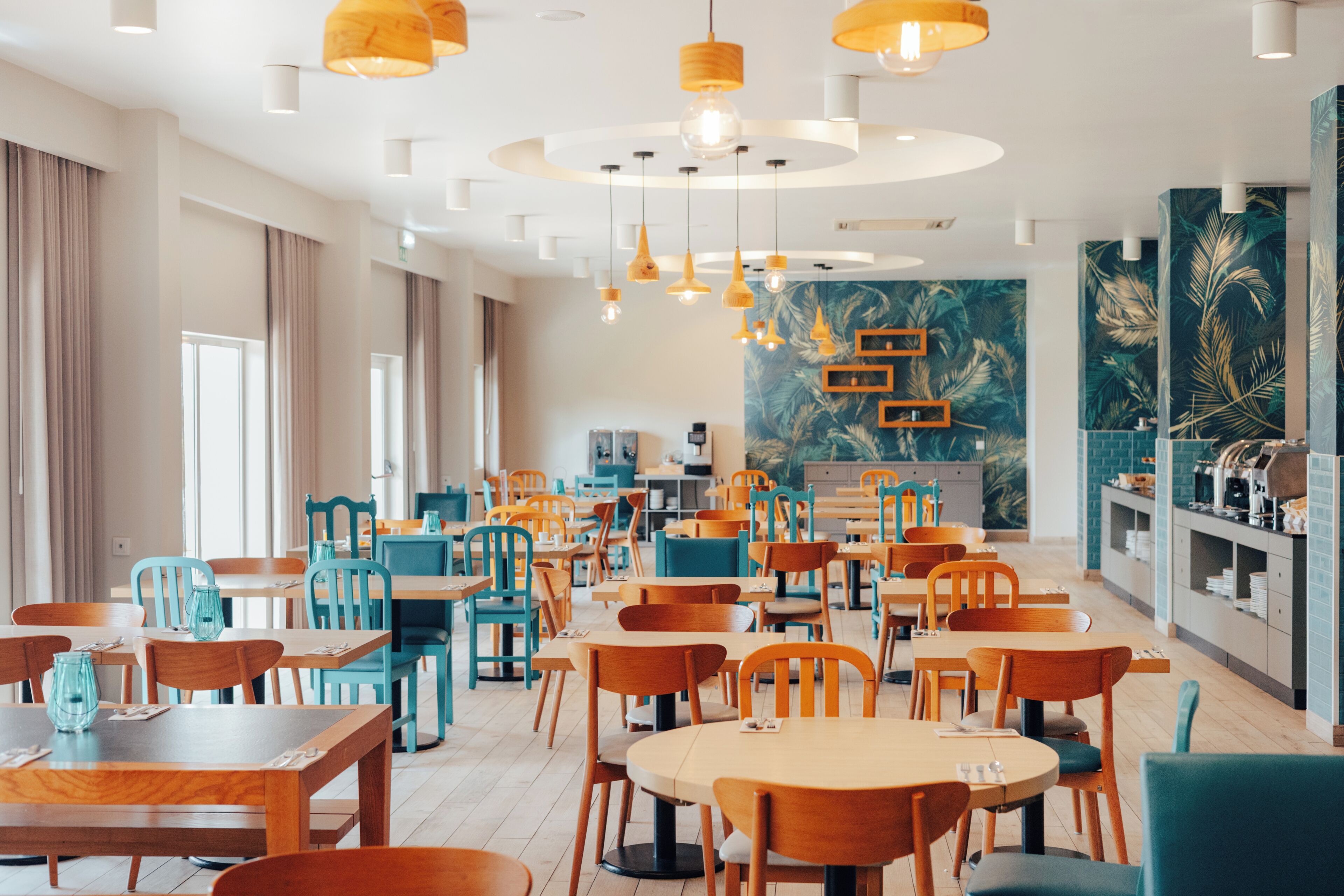 Un comedor moderno con varias mesas y sillas, iluminación en el techo y un mural temático tropical.