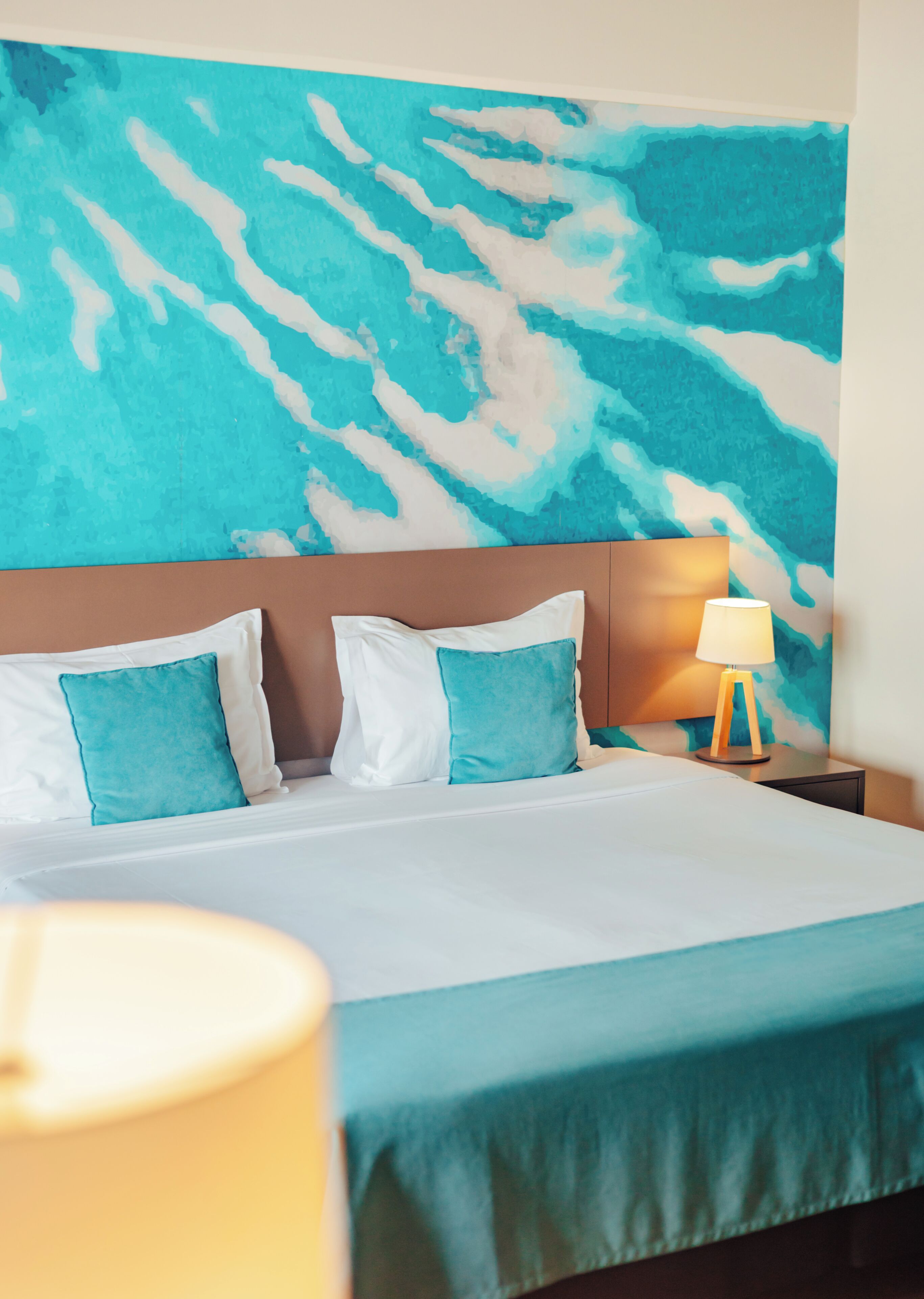 Een hotelkamer met een bed, twee kussens, een nachtkastje met een lamp en een blauw abstracte muurdecoratie.