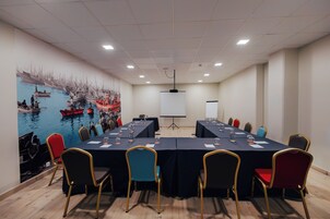 Un salón de conferencias con una mesa larga, sillas, una pantalla de proyector y un mural de barcos en la pared.