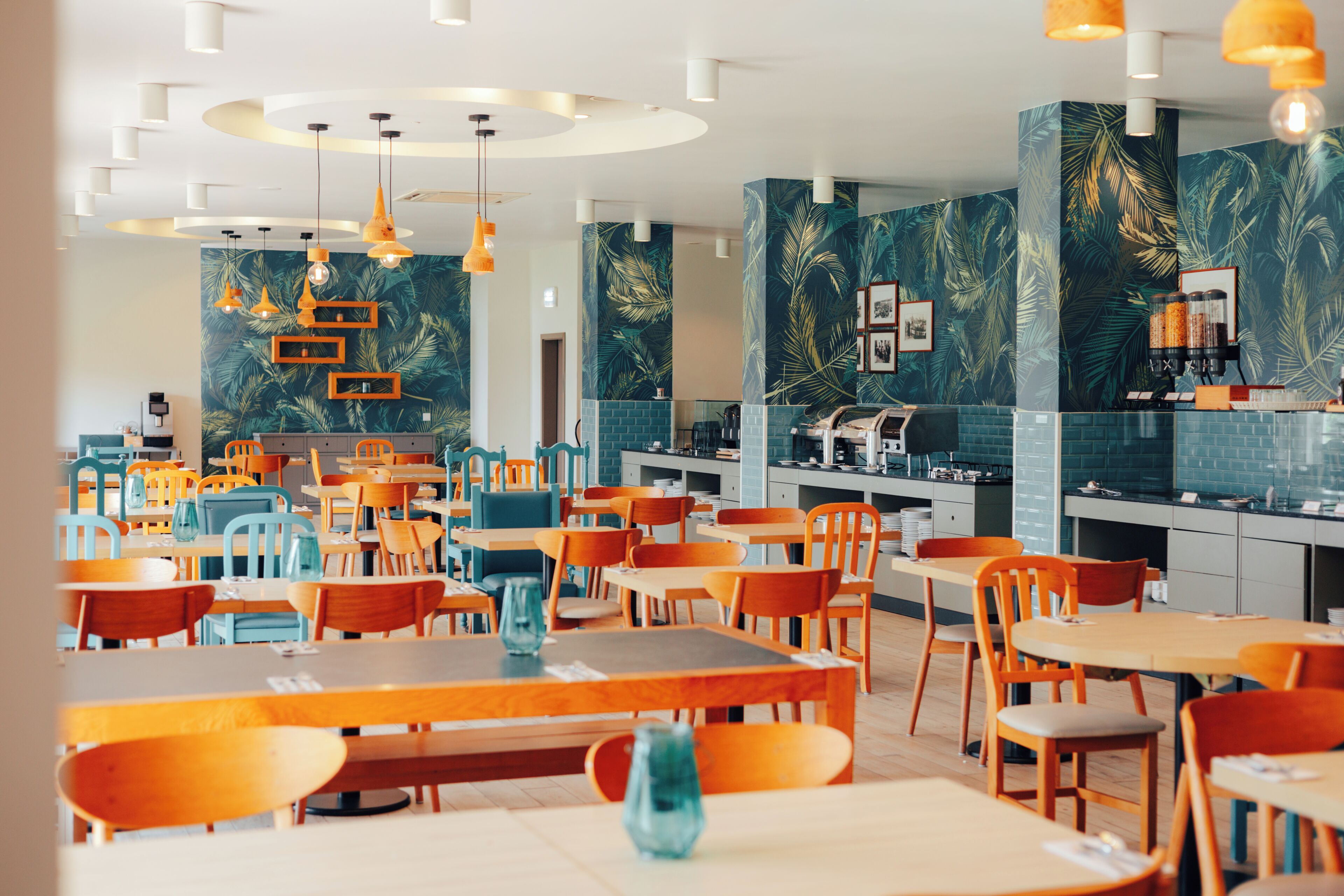 A modern restaurant interior with multiple dining tables and chairs, a kitchen area, and tropical-themed wall decorations.
