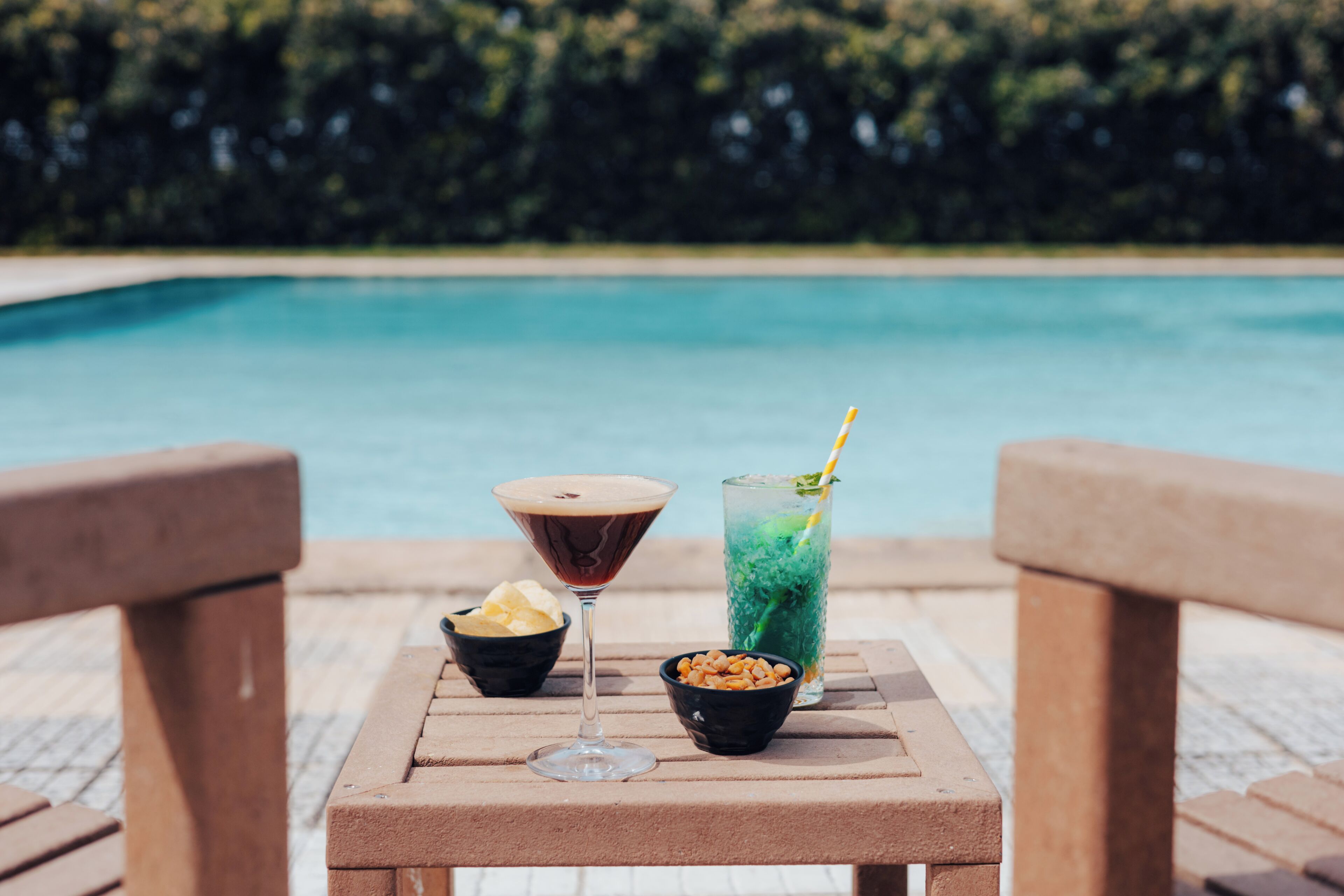 A table with a martini glass, a tall glass with a straw, and bowls of snacks by a poolside.