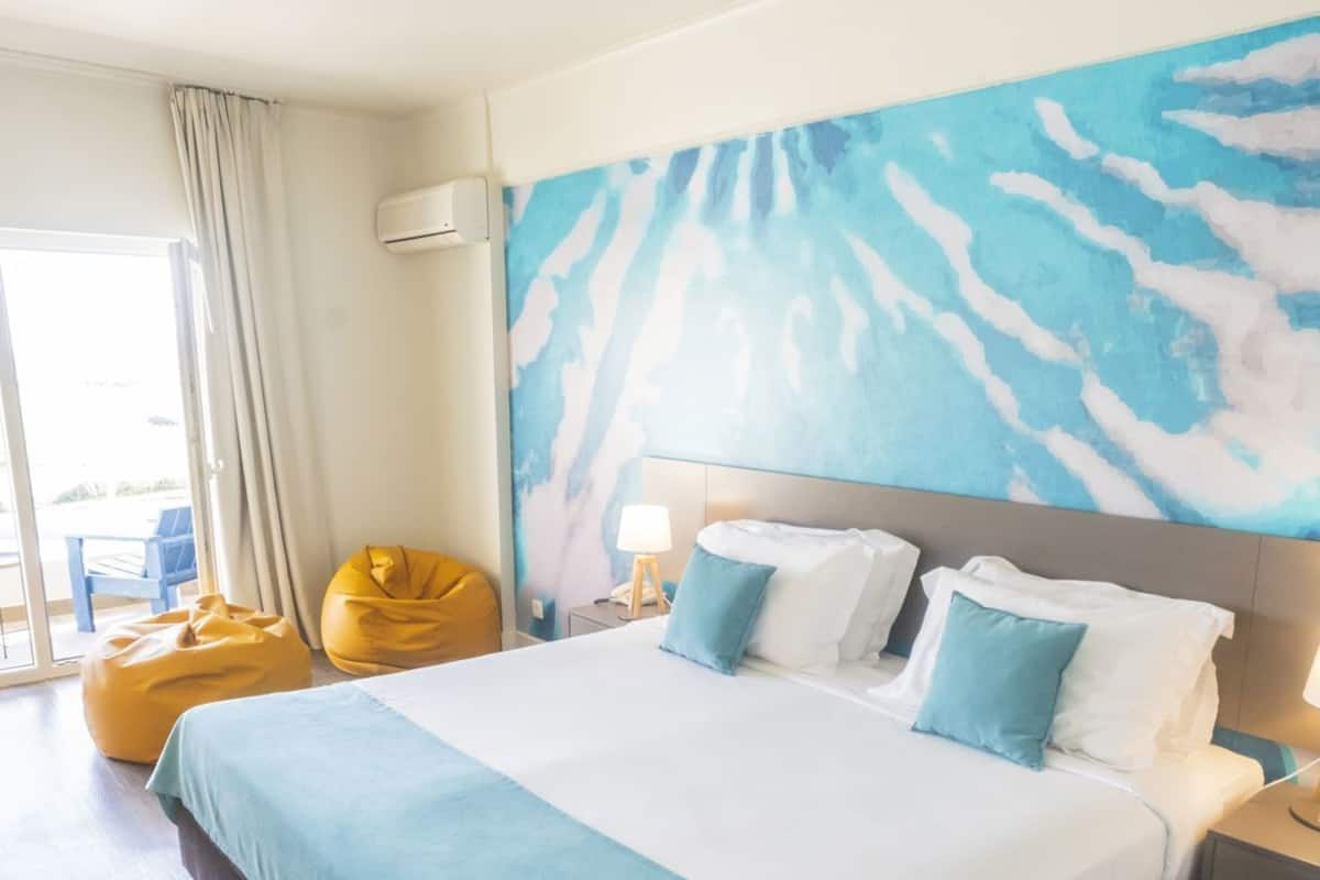 A hotel room with a large bed, a window with curtains, a wall-mounted air conditioner, and a yellow bean bag chair.