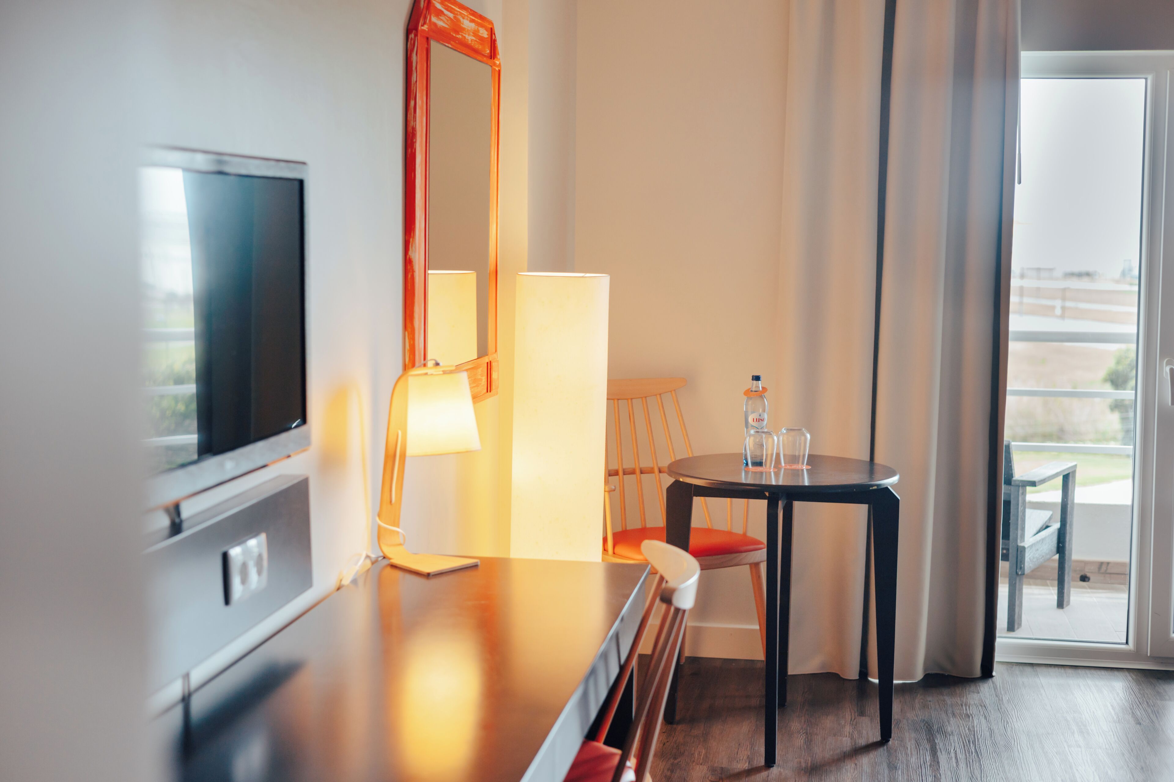 Ett hotellrum med en tv, ett bord med en stol, en lampa och en flaska på bordet.