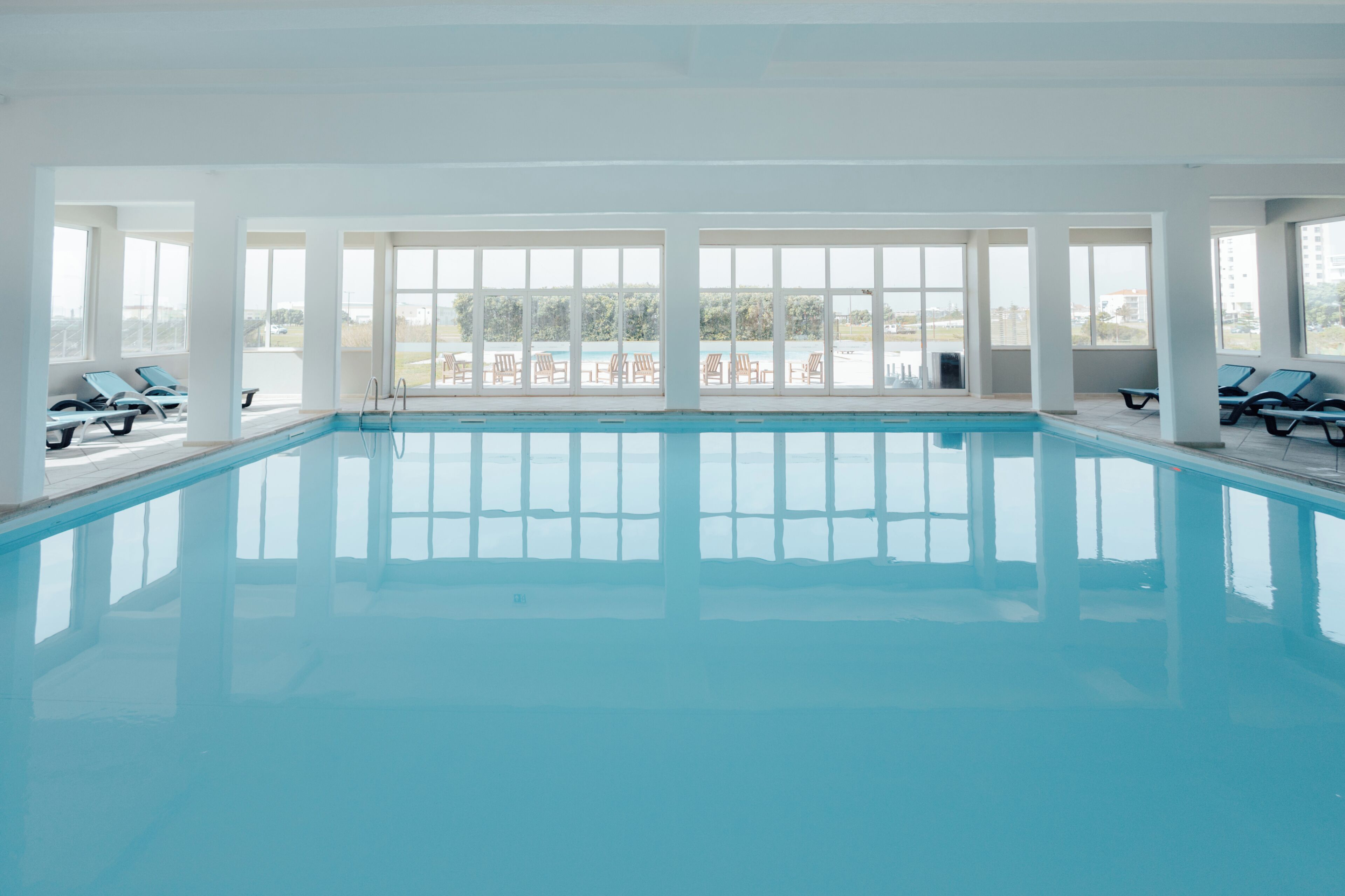 Una piscina interior con paredes de vidrio transparente, sillones de descanso y vistas al exterior.