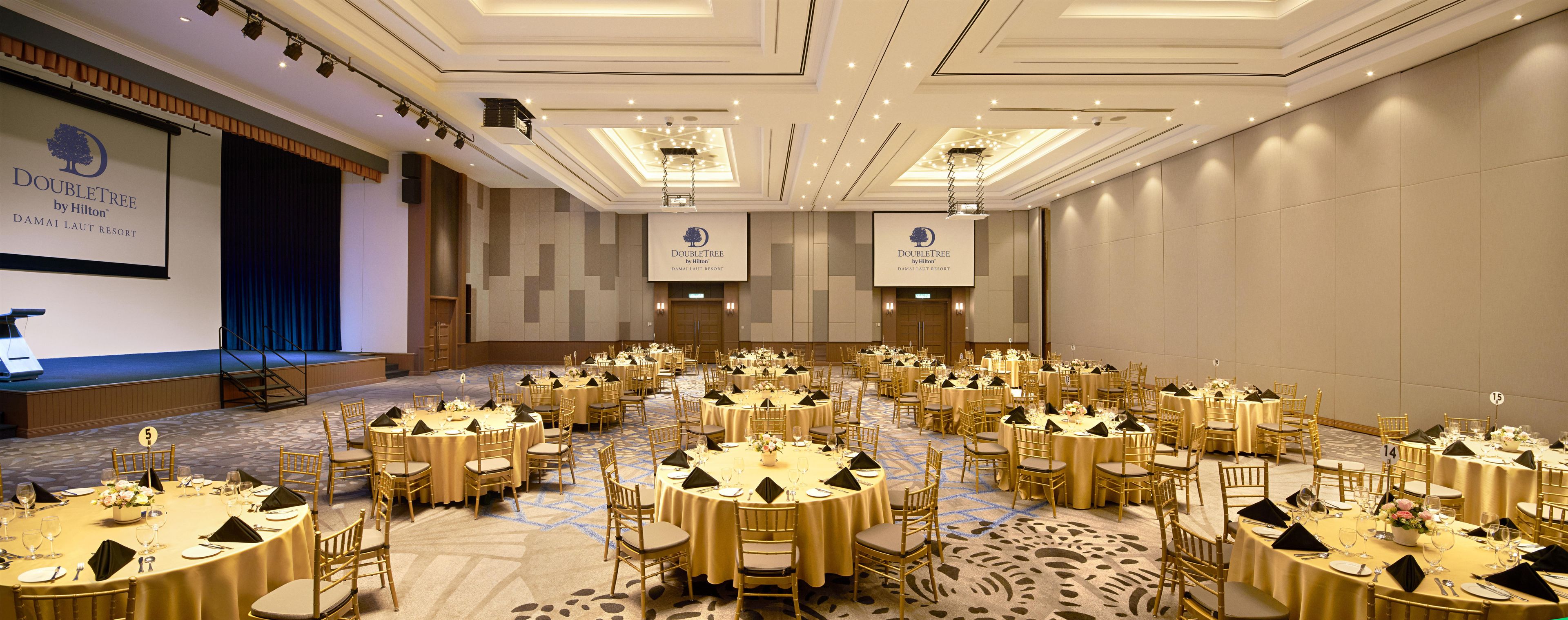 A large conference room with multiple round tables, each set with gold chairs and white tablecloths, at the DoubleTree by Hilton DAMAI LAUT RESORT.