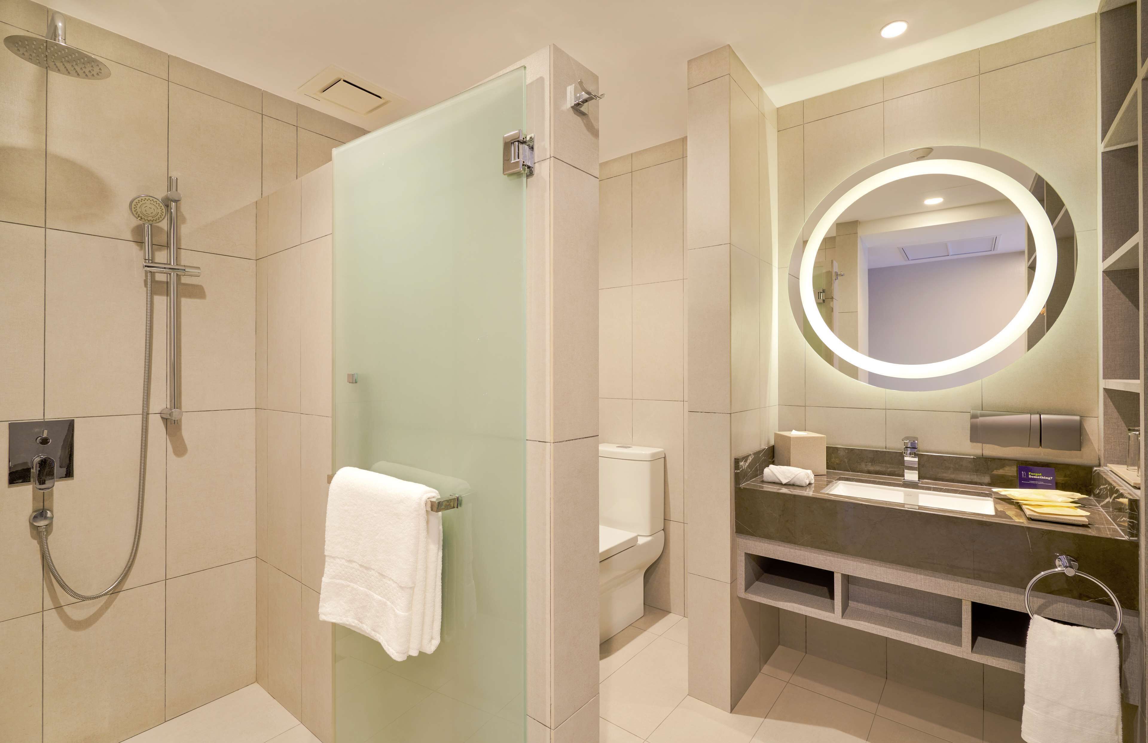 Un baño moderno con ducha envuelta en vidrio, un espejo redondo con iluminación integrada, un tocador con lavamanos y un inodoro.