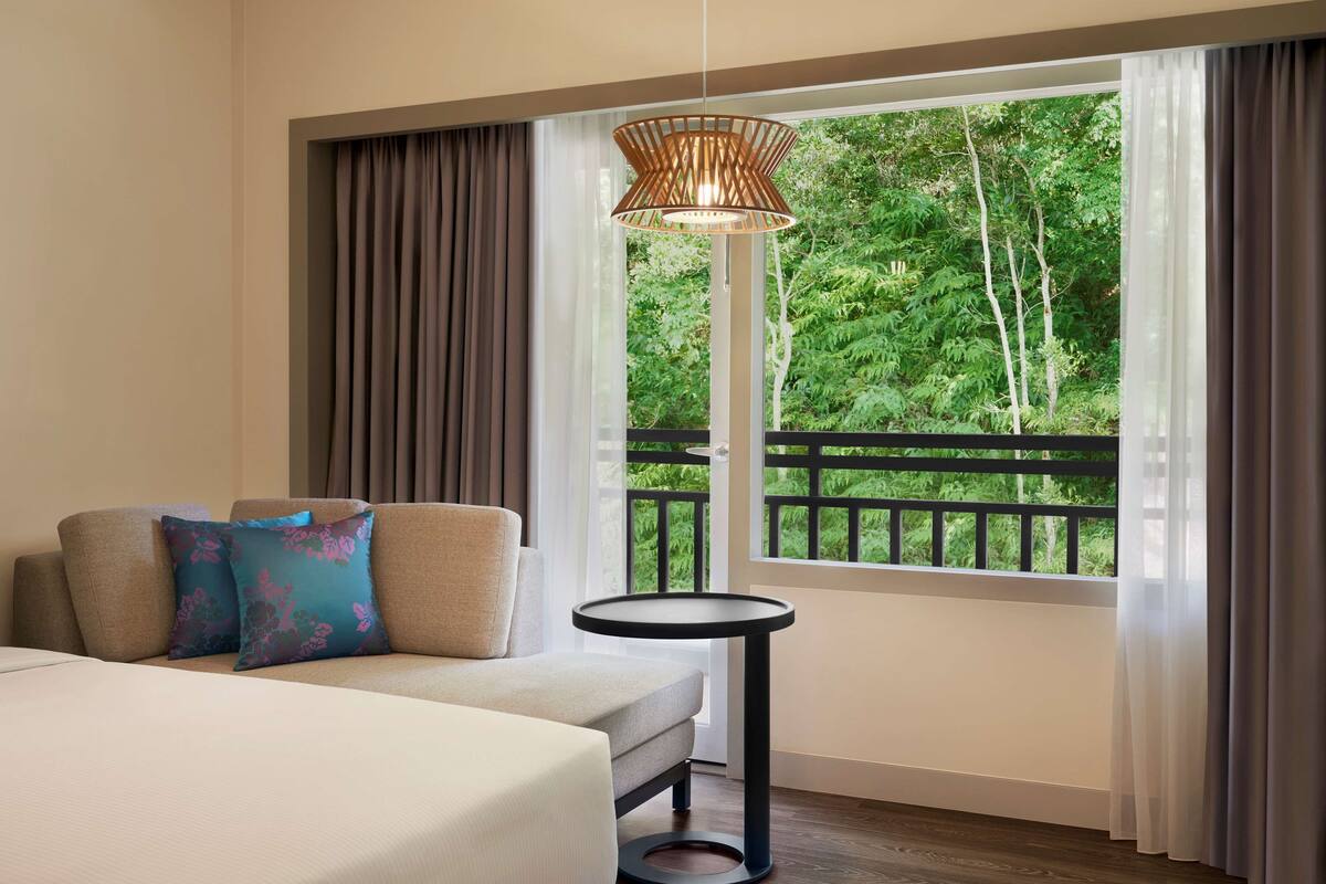 Ein Hotelzimmer mit einer Couch, Kissen, einem kleinen Tisch und einem großen Fenster mit Blick auf Grünflächen.