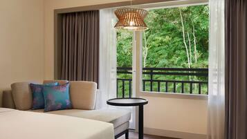 Quarto de hotel com sofá, almofadas, uma mesa pequena e uma grande janela com vista para áreas verdes.