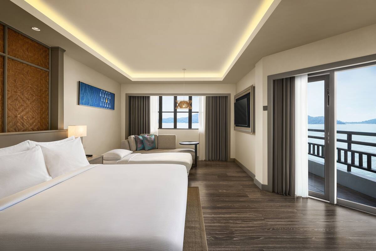 Ein Hotelzimmer mit einem großen Bett, einer Couch, einem kleinen Tisch und einem Fernseher. Ein Balkon bietet Blick aufs Meer.