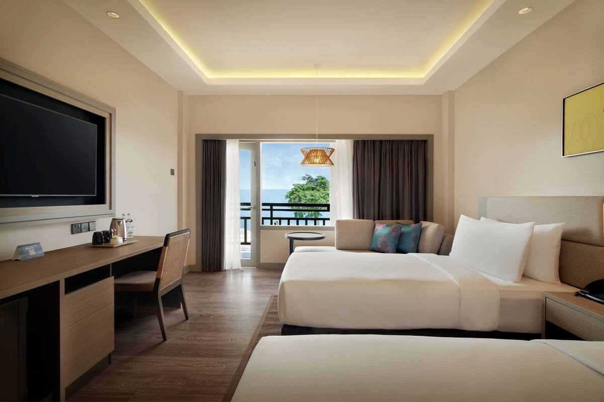 Ein Hotelzimmer mit einem großen Bett, einem Schreibtisch mit Flachbildfernseher, einem kleinen Tisch und einem Balkon mit Meerblick.