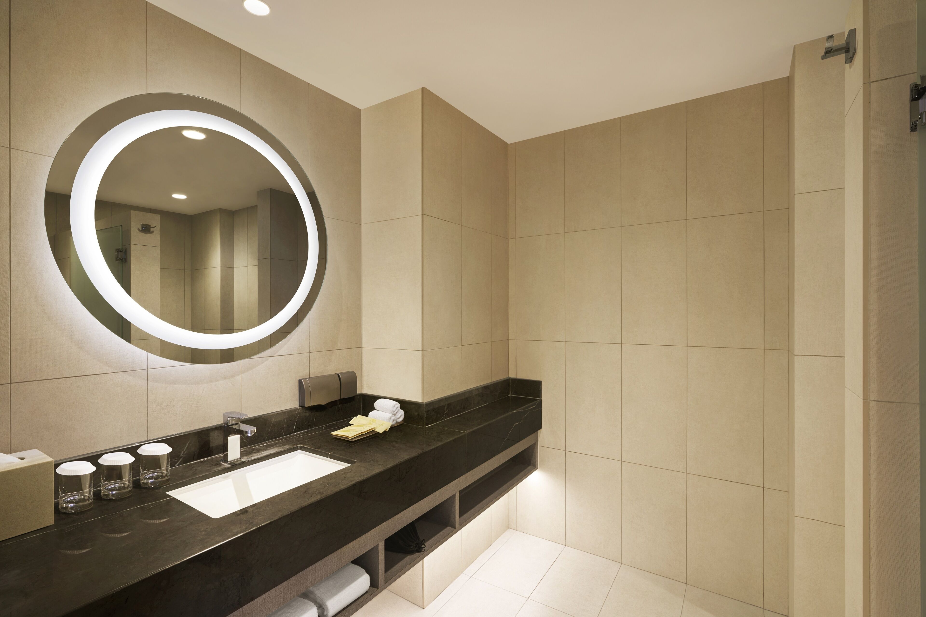 Un baño moderno con un espejo grande, una encimera de mármol oscuro, un lavabo blanco y paredes cubiertas de baldosas beige.