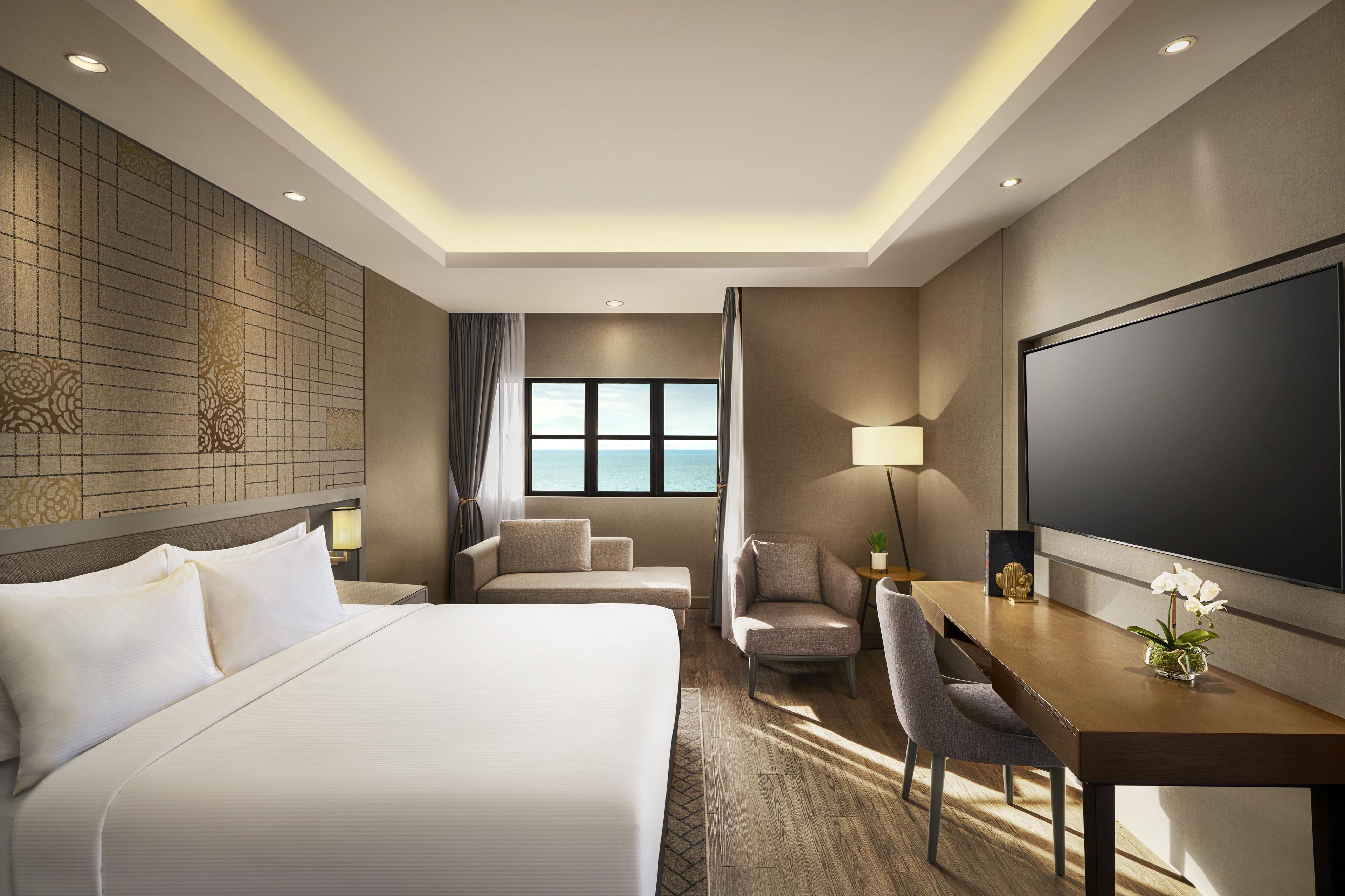 A hotel room with a bed, a desk, a chair, a television, and a window with a view of the sea.