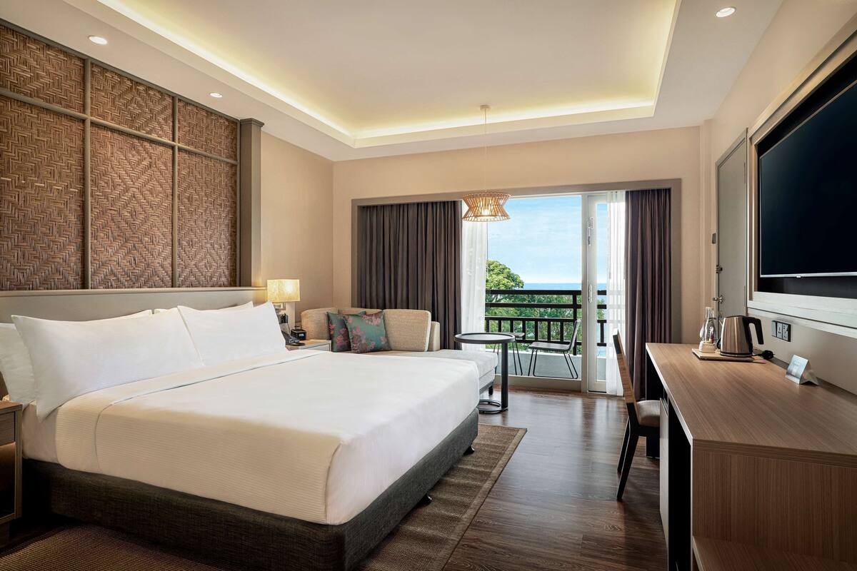 Ein Hotelzimmer mit einem großen Bett, einem Schreibtisch mit Fernseher, einem Balkon mit Blick und einer dekorativen Wandverkleidung.
